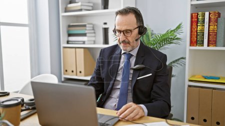 Photo for Hispanic middle-aged man with grey hair, focused on business work in the office, professionally speaking to a customer, providing aid through laptop. concentrated, successful worker using headset - Royalty Free Image