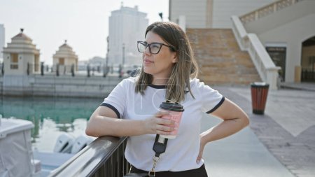 Téléchargez les photos : Une jeune femme hispanique adulte profite d'une pause café par la ligne d'horizon doha moderne dans qatar. - en image libre de droit