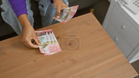 African woman counting new zealand currency indoors at a wooden table, with a focus on her hands and the money.
