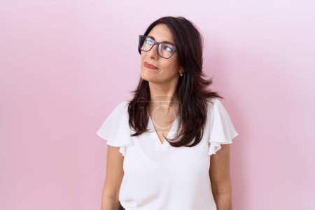 Foto de Mujer hispana de mediana edad vistiendo camiseta blanca casual y gafas sonriendo mirando hacia un lado y mirando hacia otro pensando. - Imagen libre de derechos