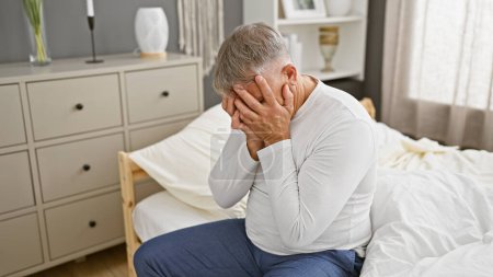 Foto de Un anciano angustiado se sienta sosteniendo la cabeza en un dormitorio moderno, transmitiendo preocupaciones, enfermedades o problemas de salud mental.. - Imagen libre de derechos