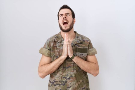 Téléchargez les photos : Jeune homme hispanique portant uniforme de l'armée de camouflage mendiant et priant avec les mains ainsi que l'expression de l'espoir sur le visage très émotionnel et inquiet. mendier. - en image libre de droit