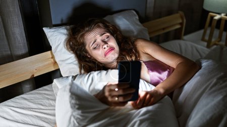 Photo for Young caucasian woman looks at smartphone with sadness in a dimly lit bedroom - Royalty Free Image