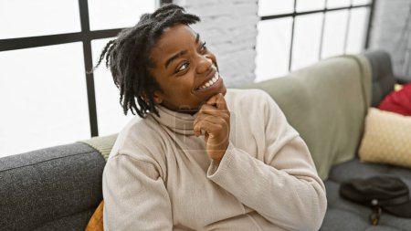 Téléchargez les photos : Une jeune femme noire souriante avec des dreadlocks portant un col roulé à la maison sur un canapé. - en image libre de droit