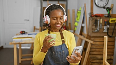 Téléchargez les photos : Femme afro-américaine profitant d'une pause café et smartphone dans un atelier de travail du bois. - en image libre de droit