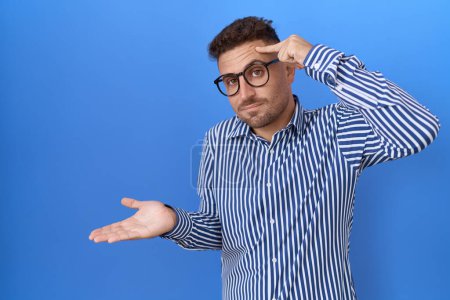 Photo for Hispanic man with beard wearing glasses confused and annoyed with open palm showing copy space and pointing finger to forehead. think about it. - Royalty Free Image