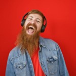 Confident young redhead guy loving his music, dancing and having fun, over an isolated red background while listening in casual attire, oozing positivity and happiness