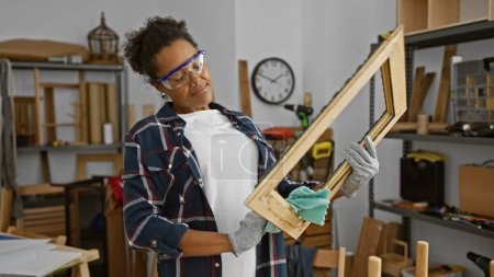 Adult african american woman working in a woodshop studio, examining a wooden frame with safety goggles on.