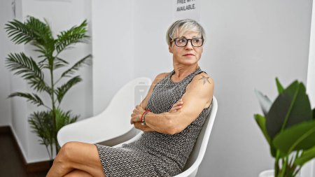 Mature hispanic woman with grey short hair sitting with arms crossed in a white indoor room