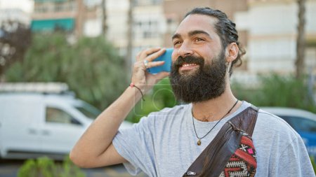 Photo for Handsome hispanic man with beard smiling while talking on smartphone on city street - Royalty Free Image
