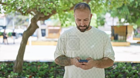 Photo for Handsome, young caucasian man with a serious expression, deeply engrossed in his smartphone, standing in the sunny, green park, connected to his online world while enjoying a relaxed, casual lifestyle - Royalty Free Image