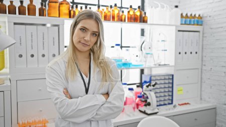 Photo for Serious-faced, beautiful blonde, young woman scientist standing, arms crossed, deep in concentration in bustling lab, amidst her medical experiment, captivating world of biology analysis. - Royalty Free Image