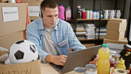 A focused young man with a headset volunteers at a charity donation center, sorting items while using a laptop.