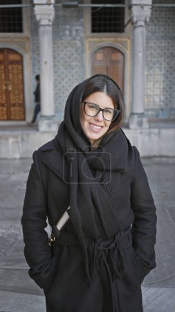 Téléchargez les photos : Une jeune femme souriante, vêtue d'un élégant manteau noir et d'une écharpe, pose devant l'architecture ornée du palais historique topkapi à Istanbul, Turquie. - en image libre de droit
