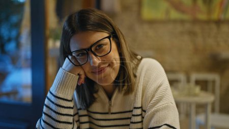 Foto de Una mujer joven se relaja en el interior de una cafetería con una sonrisa atractiva, usando gafas y exudando belleza casual.. - Imagen libre de derechos