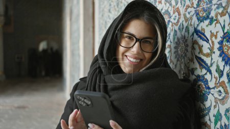 Téléchargez les photos : Une jeune femme souriante portant des lunettes et un foulard utilise un smartphone par des tuiles ornées à Istanbul. - en image libre de droit