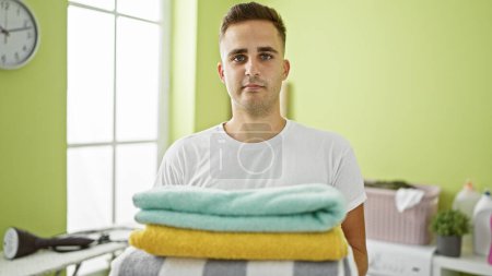 Photo for Young man standing in a laundry room holding folded clothes, with a washing machine and laundry basket in the background. - Royalty Free Image