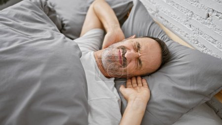 Téléchargez les photos : Épuisé, homme d'âge moyen aux cheveux gris, confortablement reposant dans le confort douillet de sa chambre, allongé, relaxant sur son lit doux, profondément endormi, échappant à la ruée matinale à l'intérieur. - en image libre de droit