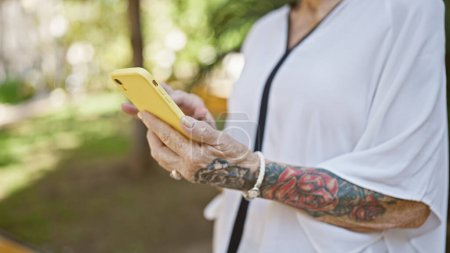 Photo for Senior woman in city park engages with technology, touching, typing & texting messages on her smartphone - Royalty Free Image