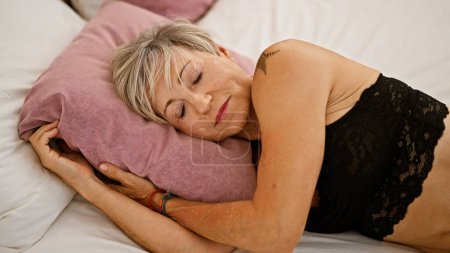A serene mature woman rests in a bedroom, embracing a pillow, with grey hair and closed eyes conveying tranquility.