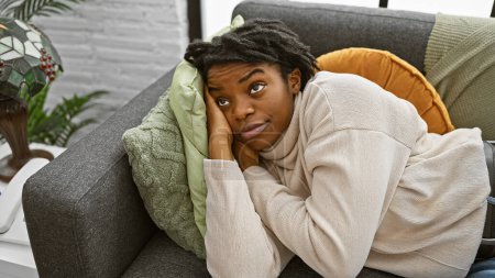 Téléchargez les photos : Une jeune femme noire contemplative avec des salons dreadlocks à la maison sur un canapé, entourée de coussins confortables. - en image libre de droit