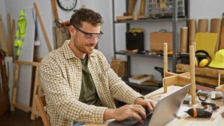 Handsome man using laptop in workshop with wood, tools, and safety gear around him.