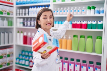 Foto de Mujer de mediana edad farmacéutico sonriendo confiado sosteniendo paquete de vitaminas en la farmacia - Imagen libre de derechos