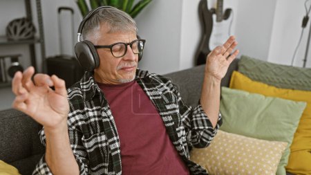 Mature man listens to music with headphones feeling relaxed and content in a cozy living room setting.