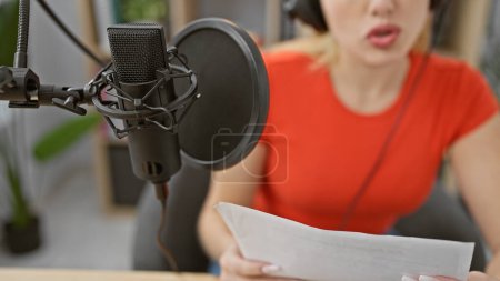 Téléchargez les photos : Une jeune femme caucasienne lit un script tout en enregistrant dans un studio de musique d'intérieur, mettant en vedette ses cheveux blonds et sa tenue rouge. - en image libre de droit
