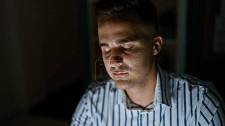 A young hispanic man with closed eyes appears to be dozing off in a dimly lit office room.