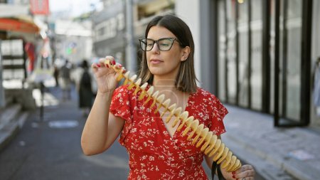 Schöne hispanische Frau, die freudig knusprige, leckere Chips auf einem Stock in der Takeshita-Straße isst, Tokyo - reisender Junk-Food-Liebhaber, Sportbrille und ein fröhliches Lächeln in Japans urbaner Stadt