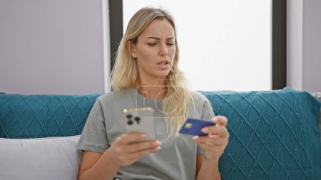 Foto de Una joven confundida examina su tarjeta de crédito mientras sostiene un teléfono inteligente, sentado en un sofá en el interior. - Imagen libre de derechos