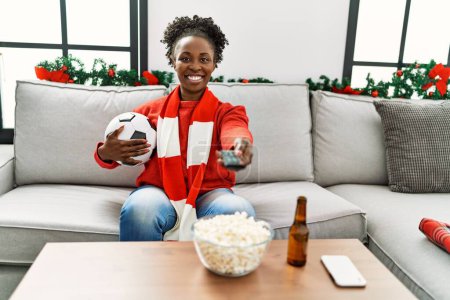Foto de Mujer afroamericana apoyando equipo de fútbol sentado en sofá en casa - Imagen libre de derechos