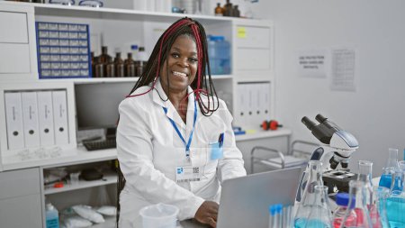 Photo for Confident african american woman, smiling widely, fully immersed in scientific research on her laptop in the bustling medical lab, beautifully braided hair catching the light as she works. - Royalty Free Image