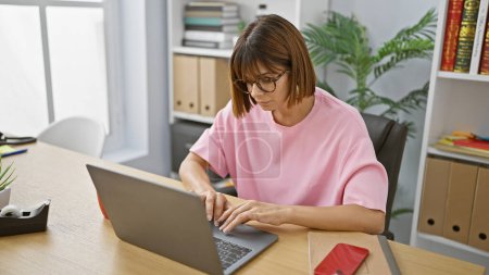 Junge, schöne, seriöse hispanische Chefin, erfolgreiche Business-Expertin, die online an ihrem Schreibtisch im Büro arbeitet, konzentriert auf ihren Computer-Laptop mit Brille