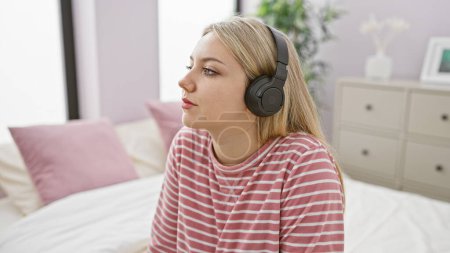 Photo for A pensive young woman wearing headphones sits in a bedroom with a striped shirt in a cozy home setting. - Royalty Free Image
