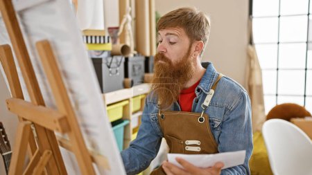 Photo for Focused redhead man intensely immersed in drawing at a bustling art studio, a young artist with beard, apron, and irish charm unraveling creativity on paper - Royalty Free Image