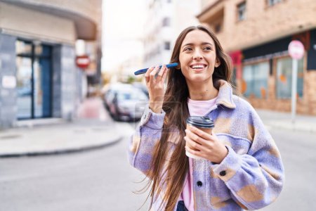 Téléchargez les photos : Jeune belle hispanique femme écoute un message vocal par smartphone boire du café dans la rue - en image libre de droit