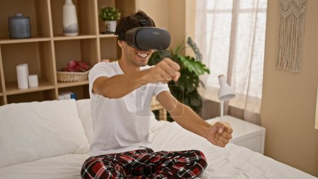 Photo for Attractive young hispanic man, gamer guy using vr glasses, confidently enjoying futuristic cyber driving game as he sits on bed, indoor home setting - Royalty Free Image