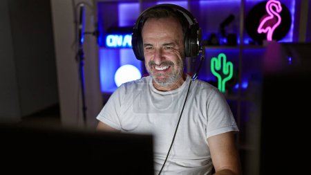 Confident middle-aged gamer with grey hair joyfully streaming his nighttime gaming session on the computer in the heart of his home gaming room