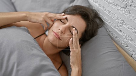 Téléchargez les photos : Belle jeune femme hispanique, stressée et souffrant de maux de tête, couchée au lit dans une chambre, reflétant son mode de vie troublé - en image libre de droit