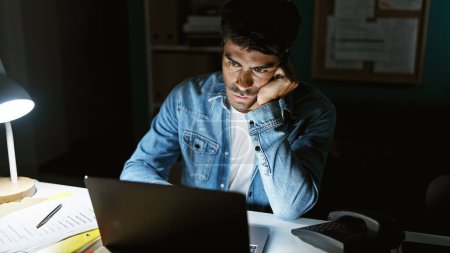 A thoughtful hispanic man with a beard works late in a dark office illuminated by a desk lamp.