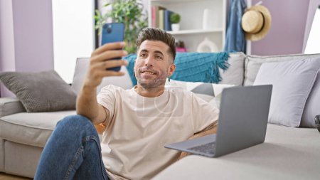 Téléchargez les photos : Jeune homme hispanique souriant profitant d'un appel vidéo décontracté sur son ordinateur portable et son smartphone, confortablement assis sur le sol de son salon à la maison - en image libre de droit