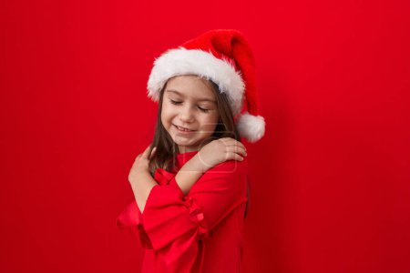 Téléchargez les photos : Adorable fille hispanique, tout sourire, se serrant dans l'amour-propre, rayonnant de bonheur sur un mur rouge isolé. revêtu d'un chapeau de Noël, respirant la confiance et les vibrations positives. - en image libre de droit