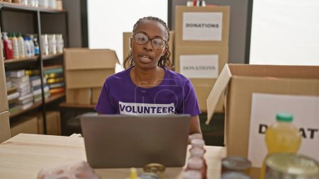 Téléchargez les photos : Femme bénévole afro-américaine travaillant sur un ordinateur portable dans un centre de dons avec des boîtes et des fournitures autour d'elle. - en image libre de droit