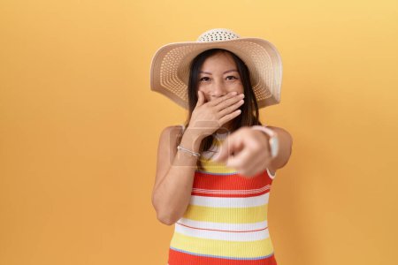Téléchargez les photos : Femme chinoise du Moyen Âge portant un chapeau d'été sur fond jaune se moquant de vous, pointant du doigt l'appareil photo avec la main sur la bouche, expression de honte - en image libre de droit