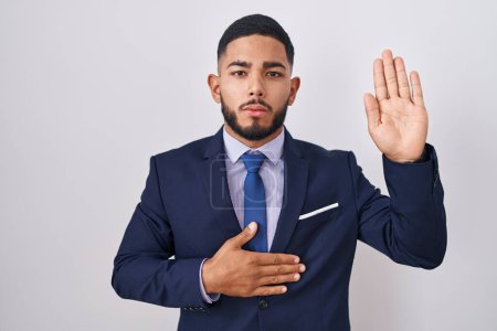 Foto de Joven hombre hispano con traje de negocios y corbata jurando con la mano en el pecho y la palma abierta, haciendo un juramento de lealtad promesa - Imagen libre de derechos