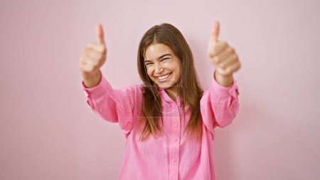 Strahlende junge hispanische Frau, die einen mit dem Daumen nach oben zeigt und selbstbewusst vor einem isolierten rosafarbenen Hintergrund steht. Ein Porträt der Positivität wie aus dem Bilderbuch!