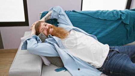 Photo for Cheerful young redhead man oozing confidence, relaxing and lying on the cozy sofa indoors, embracing positive vibes with hands on head, and flashing a handsome smile in the comfort of home - Royalty Free Image
