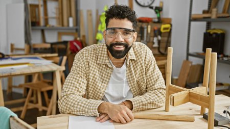 Téléchargez les photos : Homme barbu souriant en lunettes de protection assis dans un atelier de menuiserie encombré - en image libre de droit